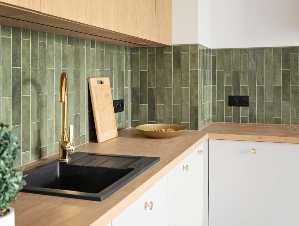 Kücheninterieur im modernen Stil mit Spülbecken und Wasserhahn, zeitgemäßen grünen Fliesen an der Wand sowie Arbeitsplatte und Möbeln aus Holz. Neue Wohnung mit stilvoller Einrichtung. Immobilien.